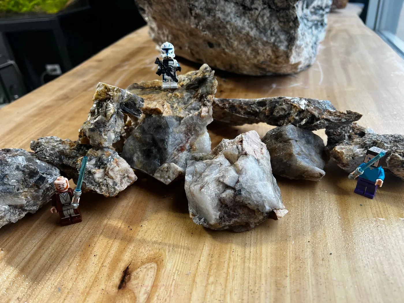 Raw pegmatite pieces with large quartz, feldspar and mica crystals on a workbench, with toy figures for scale